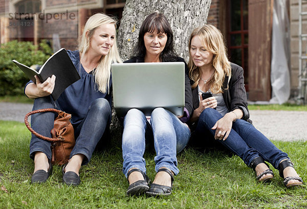 Reife Freundinnen sitzen auf dem Rasen mit Laptop