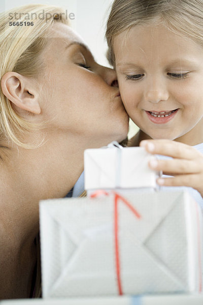 Mutter küsst die Wange der kleinen Tochter  während das Mädchen auf verpackte Geschenke schaut.