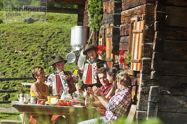 Österreich  Salzburger Land  Männer und Frauen sitzen auf einer Almhütte und hören Musikern zu.