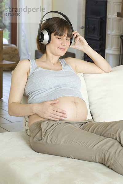 Deutschland  Nordrhein-Westfalen  Schwangere mit Kopfhörer und Sitzen auf Couch