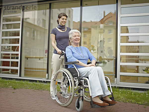 Frauen  die ältere Frauen im Rollstuhl außerhalb des Pflegeheims schieben.