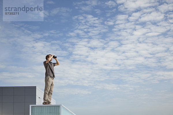 Übergroßer Mann steht auf dem Dach und schaut durch ein Fernglas.