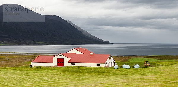 Island  Scheune auf Ackerland an der Küste mit Bergen im Hintergrund