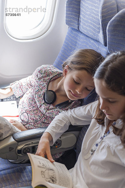 Mädchen im Flugzeug lehnt sich rüber  um sich das Buch der älteren Schwester anzusehen.