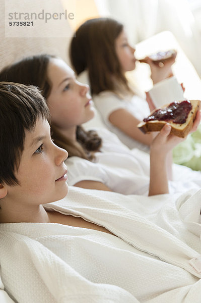 Geschwister entspannt im Bett  Mädchen essen Toasts im Hintergrund