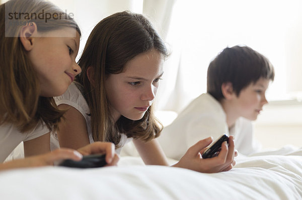 Schwestern liegen auf dem Bett und spielen Handheld-Videospiel  Junge im Hintergrund