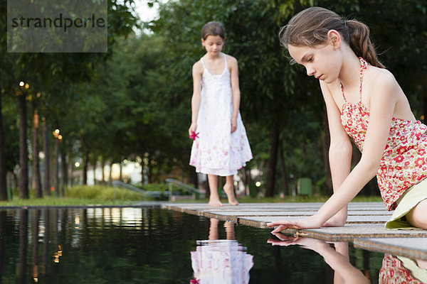 Mädchen sitzt am Teichrand und berührt die Wasseroberfläche  ein anderes Mädchen im Hintergrund