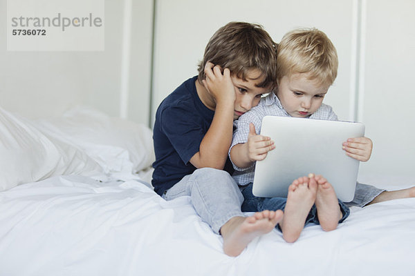 Junge Brüder sitzen auf dem Bett und schauen auf das digitale Tablett.