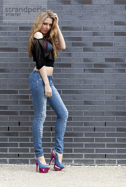 Junge Frau in zerrissenen Jeans  kurzem schwarzem Top und hohen Schuhen posiert vor Mauer