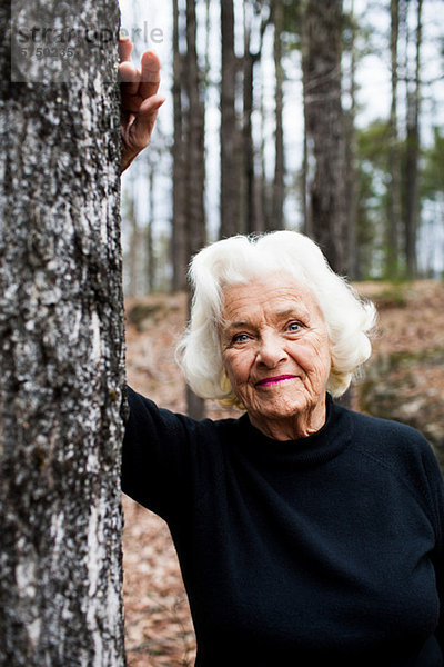 Porträt einer älteren Frau  die sich auf einen Baumstamm im Wald stützt.