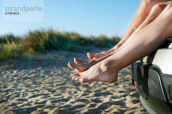 Mittleres erwachsenes Paar  das barfuß auf dem Auto am Strand sitzt.