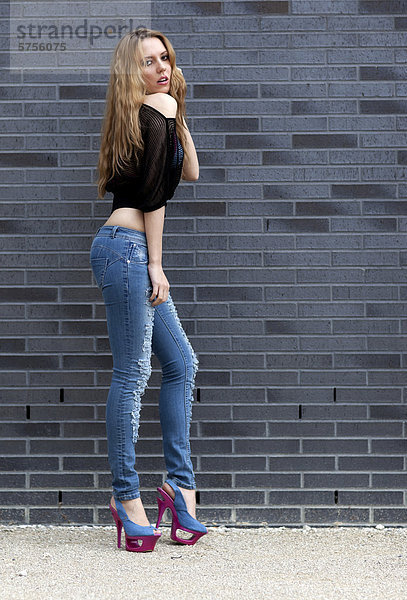 Junge Frau in zerrissenen Jeans  schwarzem Top und hohen Schuhen posiert vor Mauer