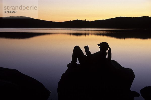 Eine junge Frau liest bei Sonnenuntergang auf einem Felsen Küstenlinie auf Stand See  an einem kühlen Herbst Nacht  Algonquin Provincial Park  Ontario  Kanada.