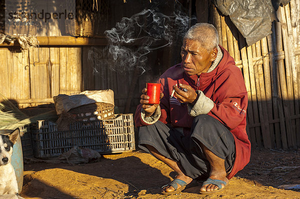 Mann raucht Zigarette und trinkt Tee vor einer Strohhütte  Dorf vom Bergvolk oder Bergstamm der Akha  ethnische Minderheit  Umgebung von Santikhiri oder Mae Salong  Chiang Rai Provinz  Nordthailand  Thailand  Asien