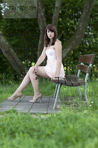 Junge Frau mit kurzem beigem Sommerkleid und hohen Schuhen sitzt draußen auf Bank