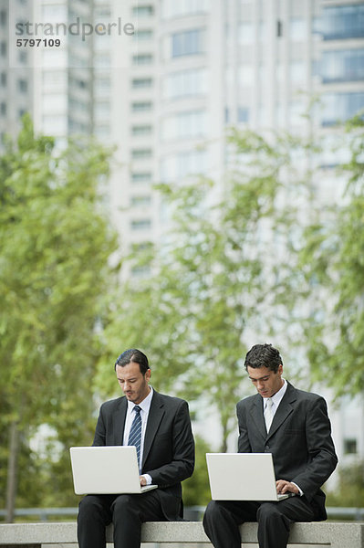 Geschäftsleute sitzen Seite an Seite und benutzen Laptops im Freien.