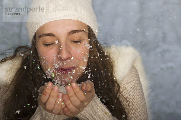 Junge Frau in Winterkleidung  bläst eine Handvoll Konfetti  Mund