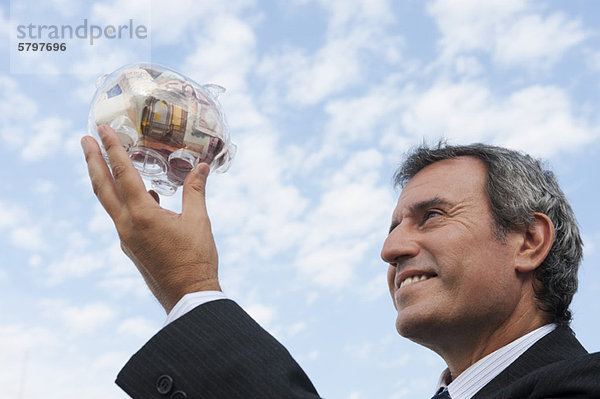Reifer Mann  der ein transparentes Sparschwein mit Euro gefüllt hält