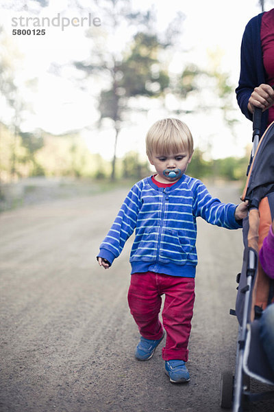 Vorschuljunge mit Schnuller im Mund  der an der Mutter entlanggeht  während er den Kinderwagen schiebt.