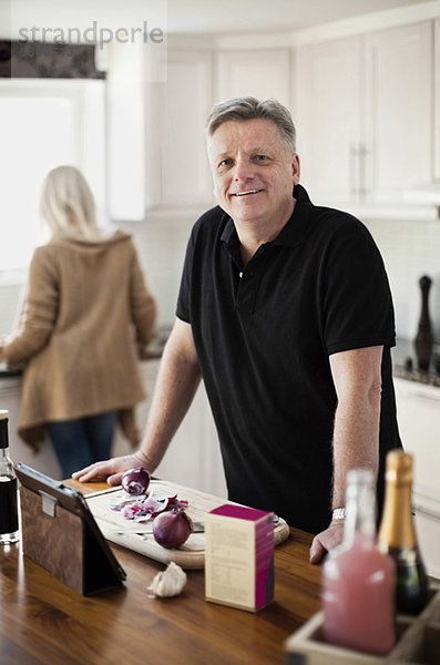 Porträt eines lächelnden reifen Mannes in der Küche mit Frau im Hintergrund