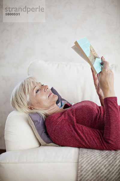 Frau liest ein Buch  während sie auf dem Sofa liegt.