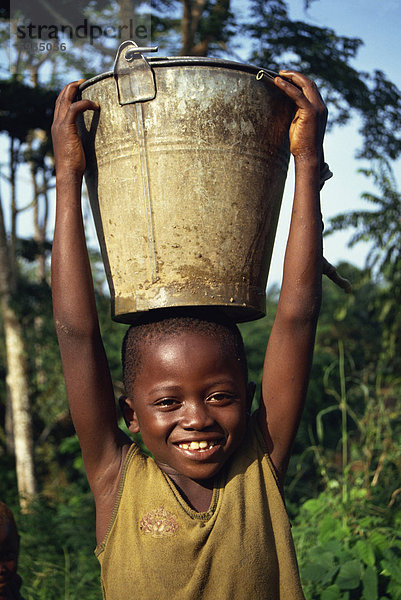 Kopf und Schultern Porträt eines jungen afrikanischen Kindes trägt einen Eimer Wasser auf dem Kopf  lächelnd und Blick auf die Kamera  Liberia  Westafrika  Afrika