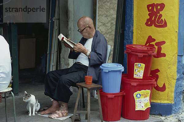 Bildnis eines älteren Mannes mit bandagierte Fuß  Brillen zu tragen und ein Buch zu lesen  sitzen mit seiner Katze neben einem Haufen von Kunststoff Lagerplätze  in Singapur  Südostasien  Asien