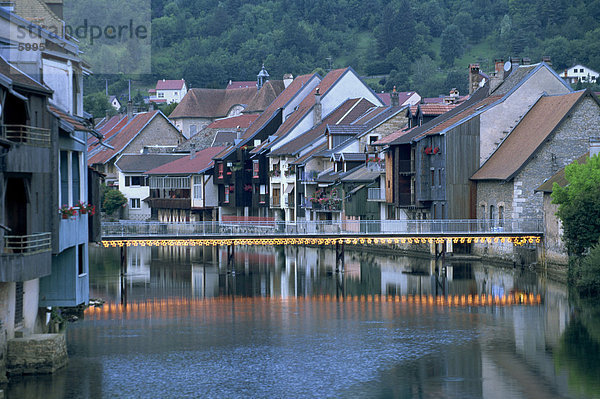 Fußgängerbrücke bei Dämmerung und Häusern entlang der Loue River  Ornans  Jura  Franche-Comte  Frankreich  Europa beleuchtet