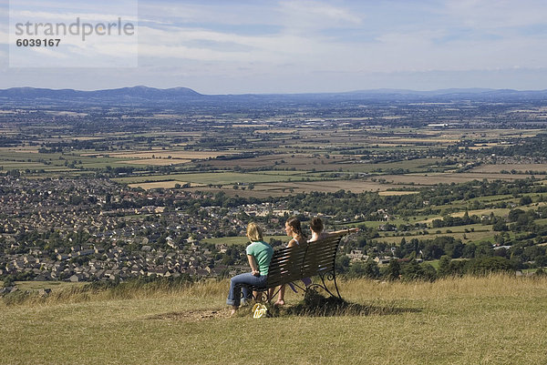 Drei Mädchen auf Bank betrachten die Ansicht über Bishops Cleeve Dorf  Cleeve Hill  The Cotswolds  Gloucestershire  England  Vereinigtes Königreich  Europa