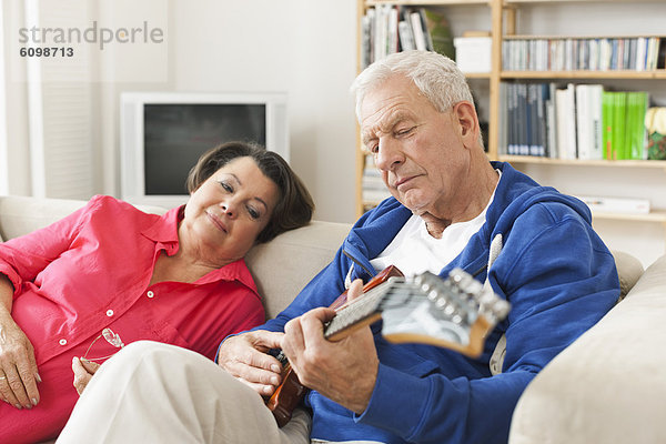 Senior Mann spielt E-Gitarre auf dem Sofa  Frau sitzt daneben