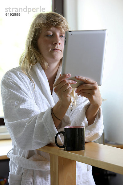 Junge Frau im Bademantel morgens mit einer Tasse Kaffee in der Küche  liest mit einem iPad  Tablet-Computer in einer Online-Zeitung