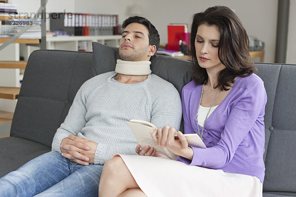 Frau liest ein Buch für ihren Mann  der an Nackenschmerzen leidet.