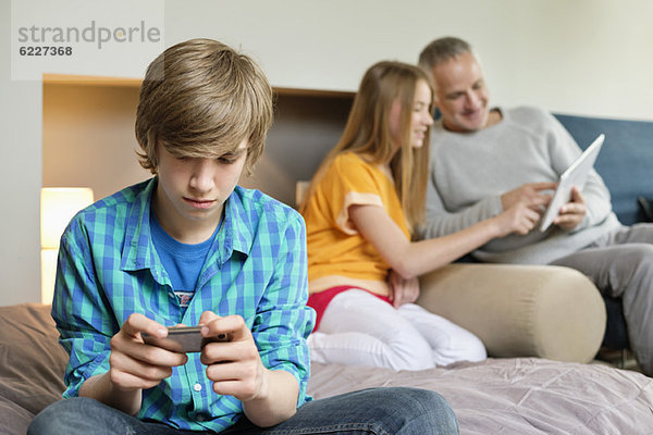 Teenager-Junge mit Handy und Vater und Schwester mit digitalem Tablett im Hintergrund