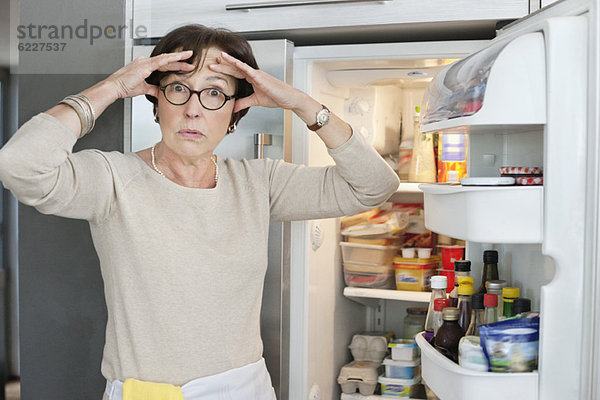 Porträt einer älteren Frau  die schockiert vor einem Kühlschrank steht.