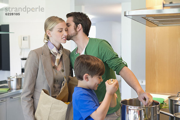 Mann  der seine Frau küsst  die vom Einkaufen zurückkehrt  und Sohn  der in der Küche kocht.