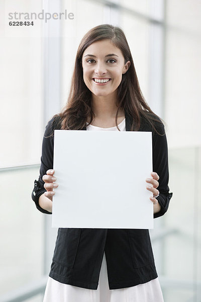 Porträt einer Geschäftsfrau  die ein leeres Plakat hält und in einem Büro lächelt.
