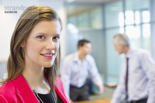 Porträt einer Geschäftsfrau  die in einem Büro lächelt und mit ihren Kollegen im Hintergrund diskutiert.