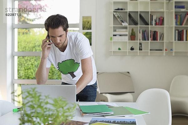 Mann  der ein Handy hält und an einem Laptop arbeitet