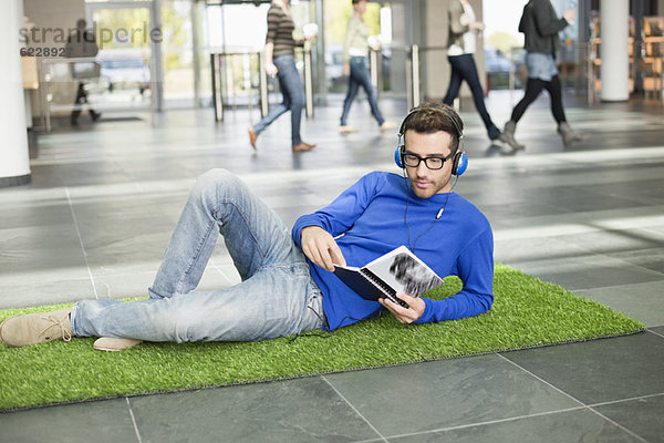 Geschäftsmann hört Musik und liest Buch  während er sich auf einer Grasmatte in einer Bürolobby ausruht.