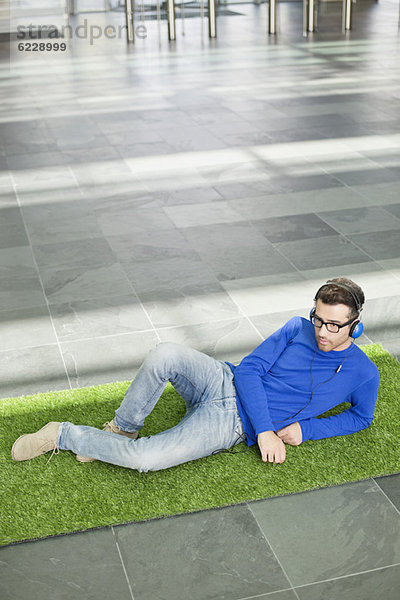 Geschäftsmann entspannt sich auf Rasen und hört Musik in einer Büro-Lobby.