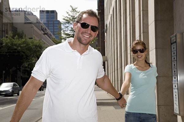 Lächelndes junges Paar mit Sonnenbrillen geht Hand in Hand auf dem Bürgersteig