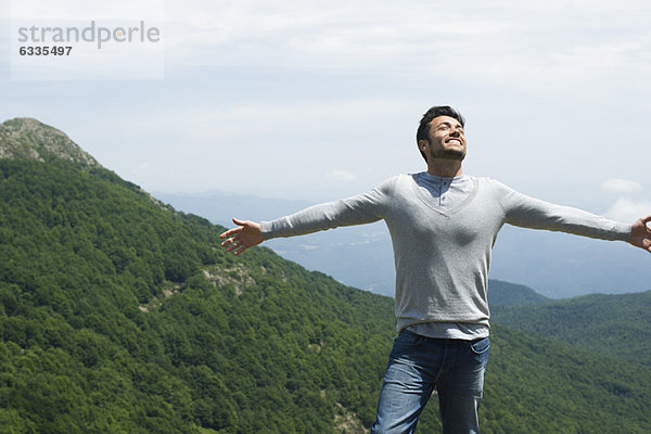 Mittelgroßer Mann mit geschlossenen Augen und ausgestreckten Armen  Berglandschaft im Hintergrund