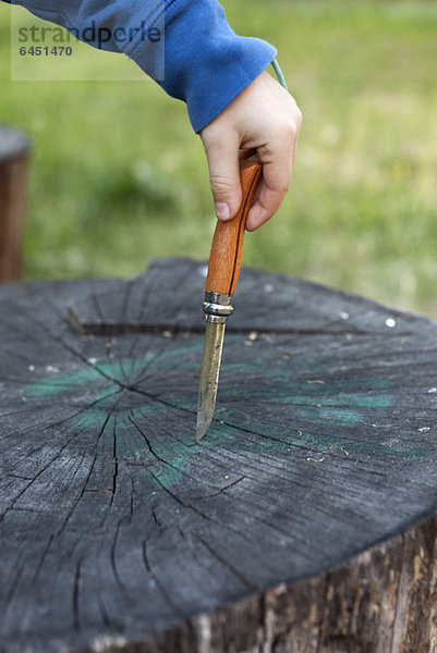 Detail eines Jungen  der ein Taschenmesser auf einem Baumstumpf hält.