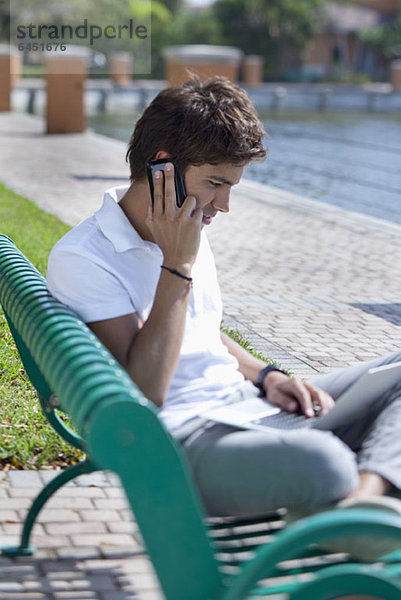 Ein Mann  der auf einer Parkbank sitzt  auf einem Handy spricht und einen Laptop benutzt.