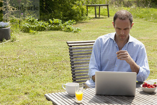 Ein Mann  der an einem Tisch in seinem Garten sitzt  frühstückt und einen Laptop benutzt.