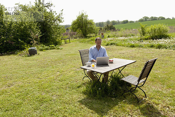 Ein Mann  der an einem Tisch in seinem Garten sitzt  frühstückt und einen Laptop benutzt.