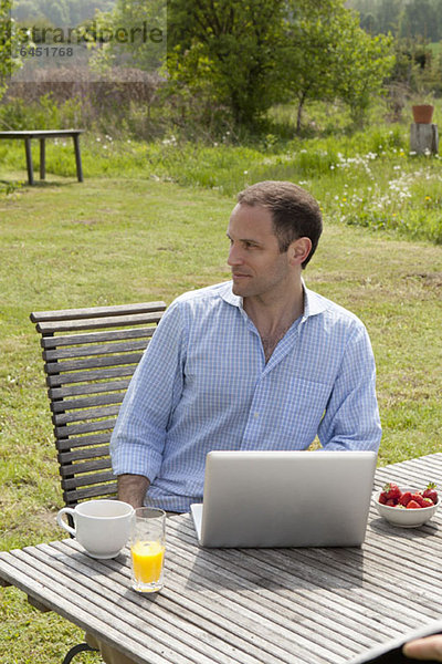 Ein Mann  der an einem Tisch in seinem Garten sitzt  frühstückt und einen Laptop benutzt.