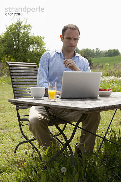 Ein Mann  der an einem Tisch in seinem Garten sitzt  frühstückt und einen Laptop benutzt.