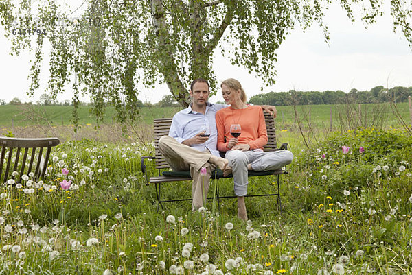 Ein Paar sitzt auf einer Bank im Garten und genießt ein Glas Rotwein.