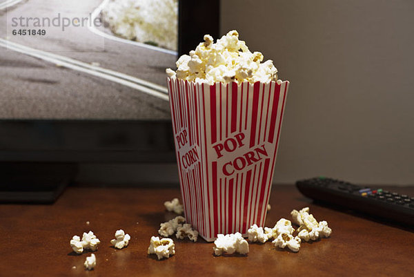 Ein rot gestreifter Karton Popcorn auf einem Tisch vor einem Flachbildfernseher
